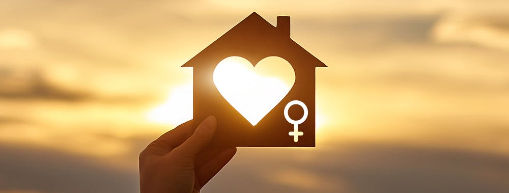 Main gauche tenant devant le soleil une silhouette de maison en bois avec découpe en forme de coeur au centre et le symbole de la femme à droite.